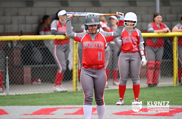 Young softball batter in red uniform (number 11) raises her bat at home plate, teammates in the dugout behind a chain-link fence.