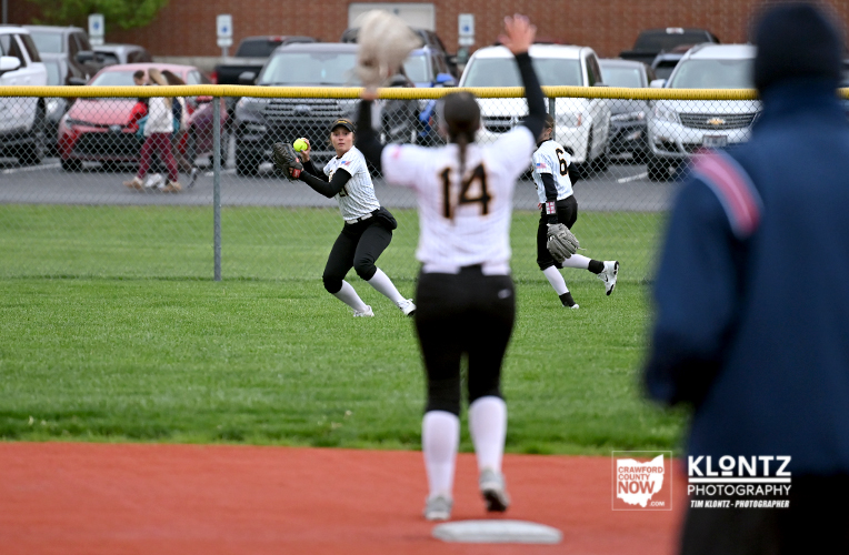 Youth softball game on a grassy field; players in white uniforms, a fielder catches the ball near the outfield fence.