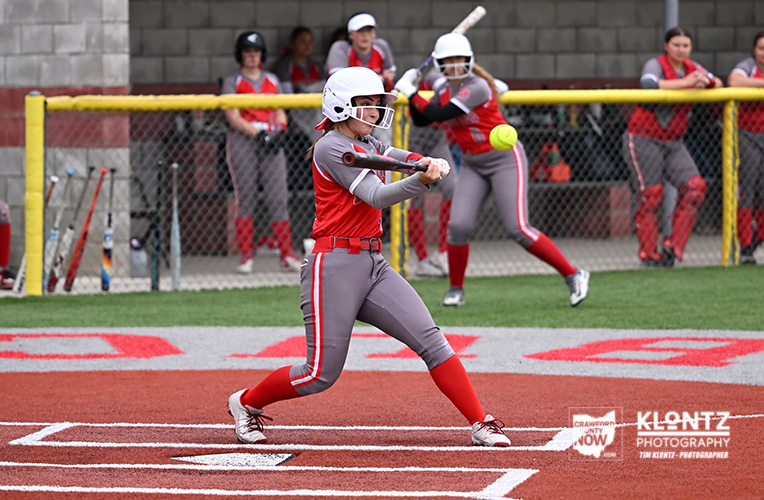 Female softball batter in red and gray uniform swings at a pitch on the field as teammates watch behind a chain-link fence.