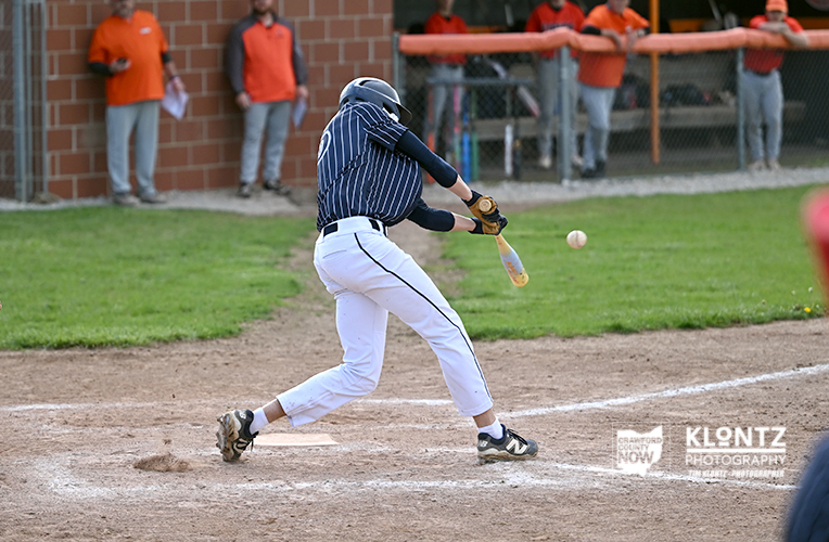Baseball batter in a navy striped jersey and white pants swings at a pitched ball at home plate, dirt and grass field visible.