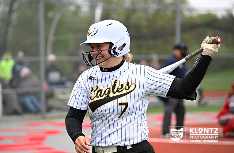 Female softball player in a white pinstripe jersey with 'Eagles' logo, wearing a helmet, swinging a bat during a game.