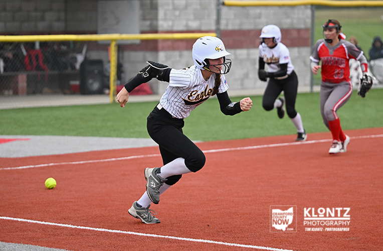 Softball player in a white helmet and striped jersey sprinting on the orange infield while a ball lies nearby and teammates chase in the background, during a game.