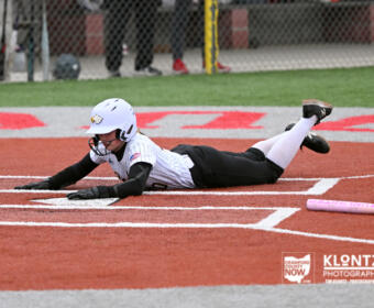 Diving baseball/softball player in a white uniform slides head-first into home plate on red dirt, a pink bat resting nearby.
