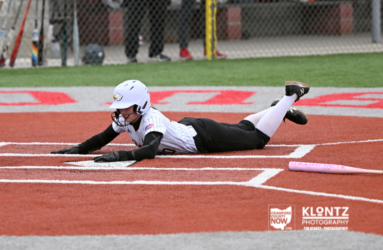Diving baseball/softball player in a white uniform slides head-first into home plate on red dirt, a pink bat resting nearby.
