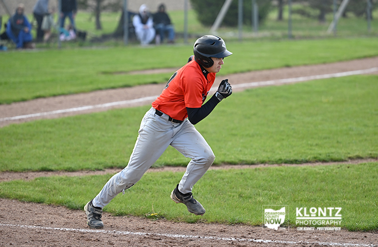 Young baseball player in an orange jersey runs along the first-base line on a grassy field, wearing a helmet and black sleeves, with spectators in the background.