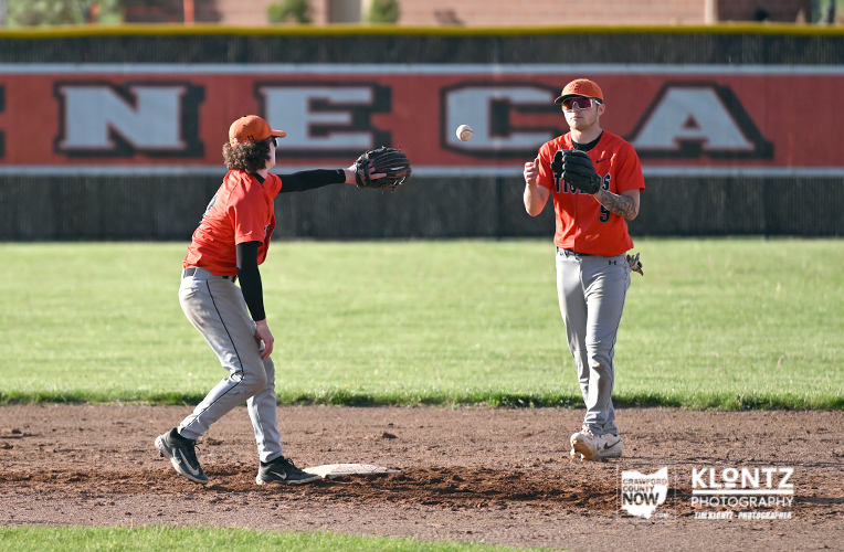 Two baseball players in orange jerseys on an infield, one reaches with a glove to field a ball while the other stands ready nearby with a glove in hand, a red stadium wall behind them.