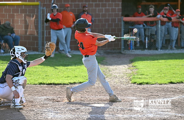 Youth baseball batter in an orange jersey swings at a pitched ball while the catcher crouches behind home plate in protective gear.