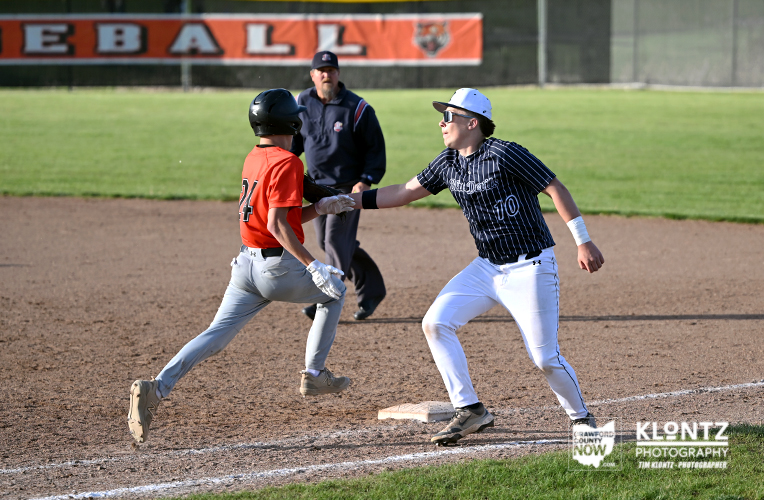 Young baseball players on a dirt infield near a base: orange-uniform runner slides toward the base as a pinstriped-fielding opponent reaches to tag, with an umpire watching in the background.