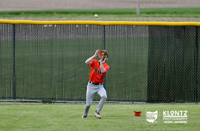 Youth baseball player in an orange jersey reaches up with a glove to catch a ball near the outfield fence.190821