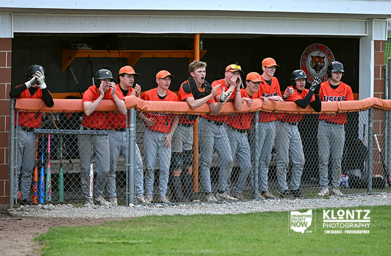 Baseball players in orange jerseys lean on the dugout railing, watching the game together.