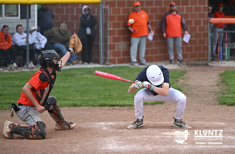 Young batter wearing a white helmet swings at a pitch as the catcher in orange gear crouches behind the plate on a dirt field.