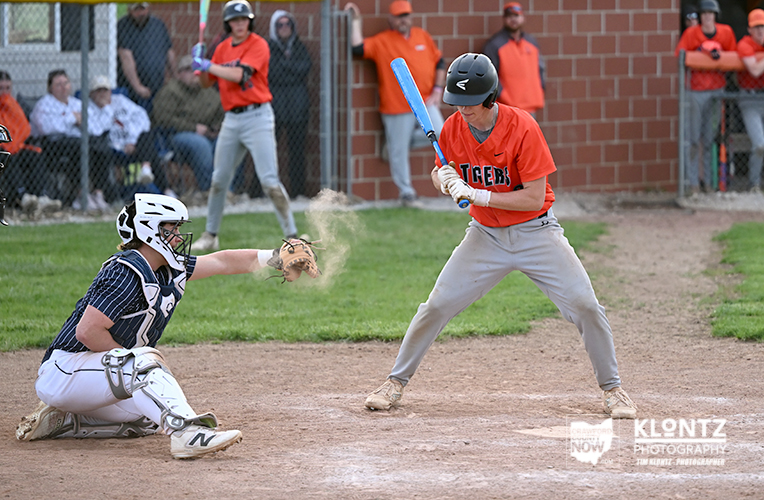 Baseball batter in an orange jersey swings at a pitch as the catcher in dark gear slides a glove toward the ball, on a dirt field with spectators watching from the dugout in the background.