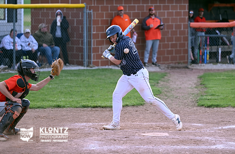 A youth baseball batter in a navy pinstripe jersey swings at a pitch at home plate, with the catcher in orange gear crouched behind him and a dirt infield.