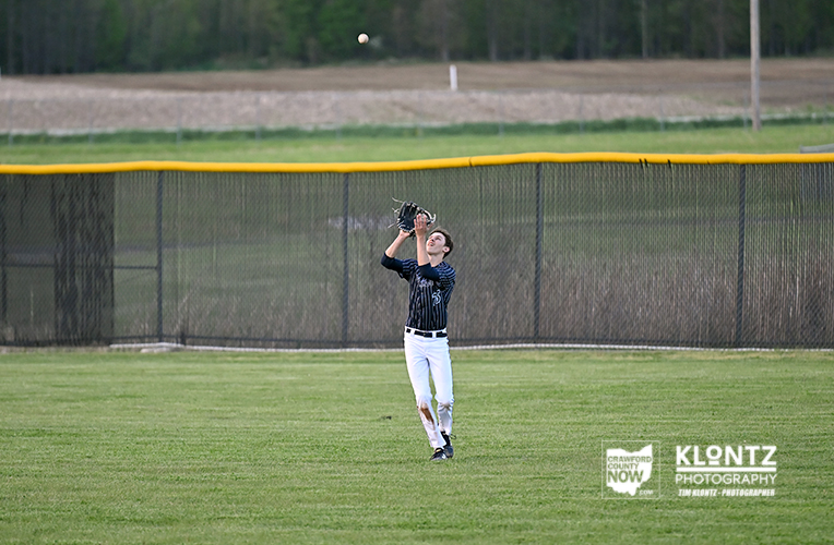Young baseball player catches a fly ball in the outfield, glove raised above head, wearing a dark striped jersey and white pants with a chain-link fence behind him.