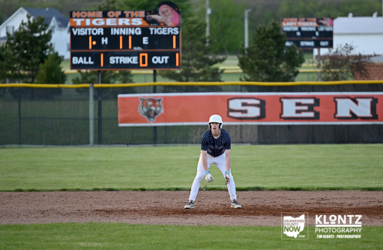 Baseball infielder wearing a white helmet and navy pinstriped jersey crouches on the infield dirt, ready to field a ball with a scoreboard in the background.