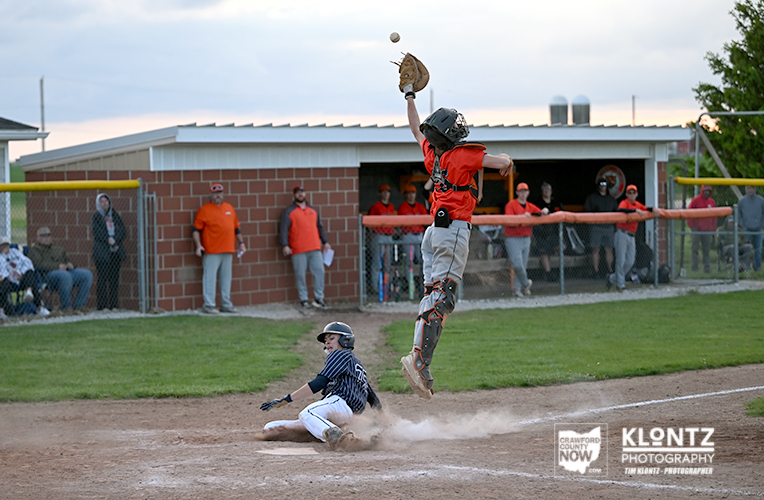 Baseball scene: a catcher in orange leaps to catch a throw as a baserunner slides into a base, kicking up dust.