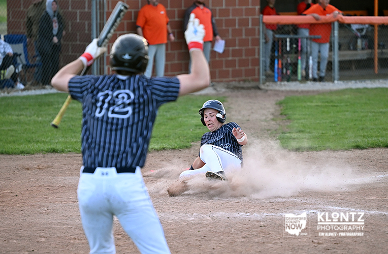 Youth baseball game: a baserunner slides into a dusty base while a fielder in a striped uniform watches from the foreground.