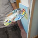 Artist holds a paint-splattered palette and brush, preparing to paint in a studio.
