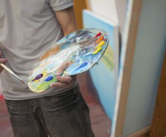 Artist holds a paint-splattered palette and brush, preparing to paint in a studio.