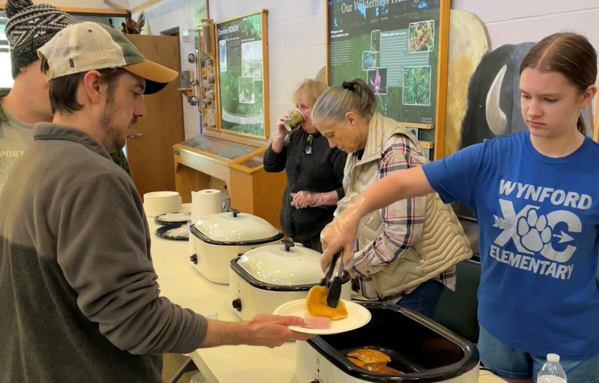 People serve pancakes from slow cookers at a community breakfast event, with volunteers and attendees lining up at a table.",