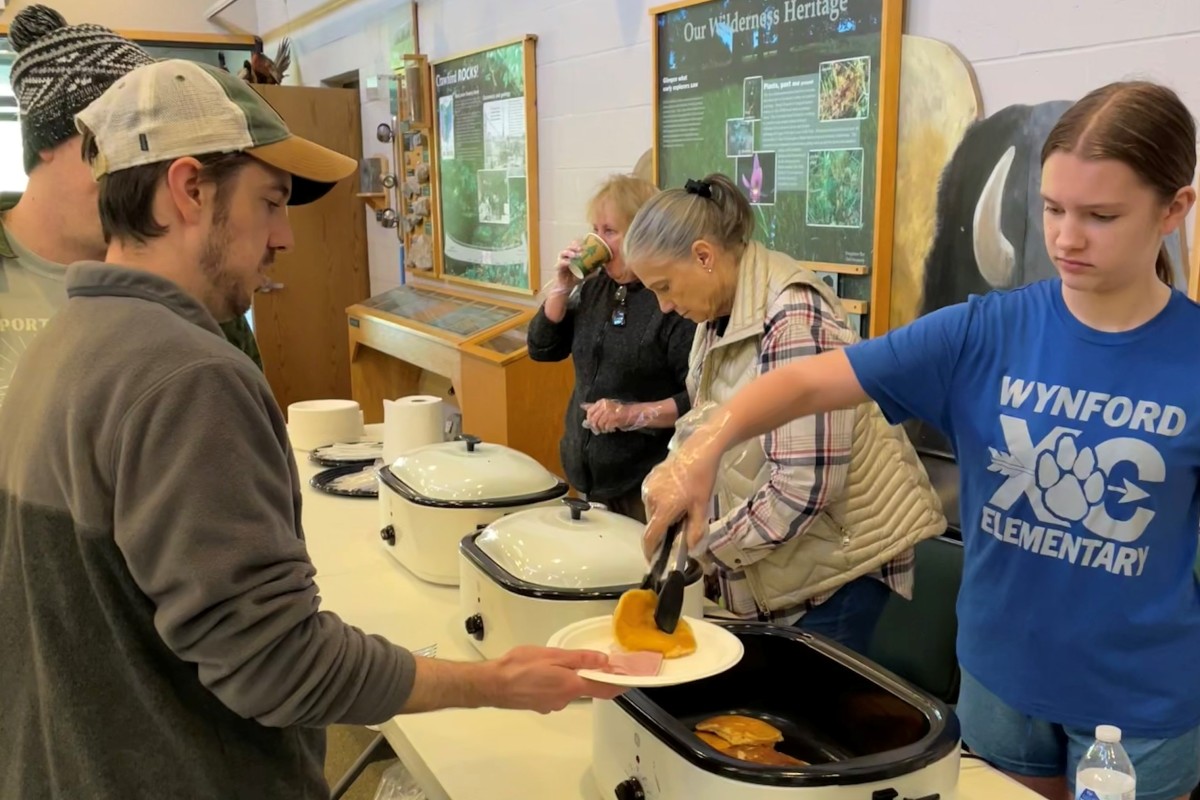People serve pancakes from slow cookers at a community breakfast event, with volunteers and attendees lining up at a table.",