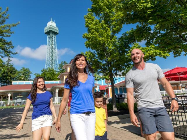 A family of four walking outdoors on a sunny day, with a tall lattice tower in the background and green trees around.