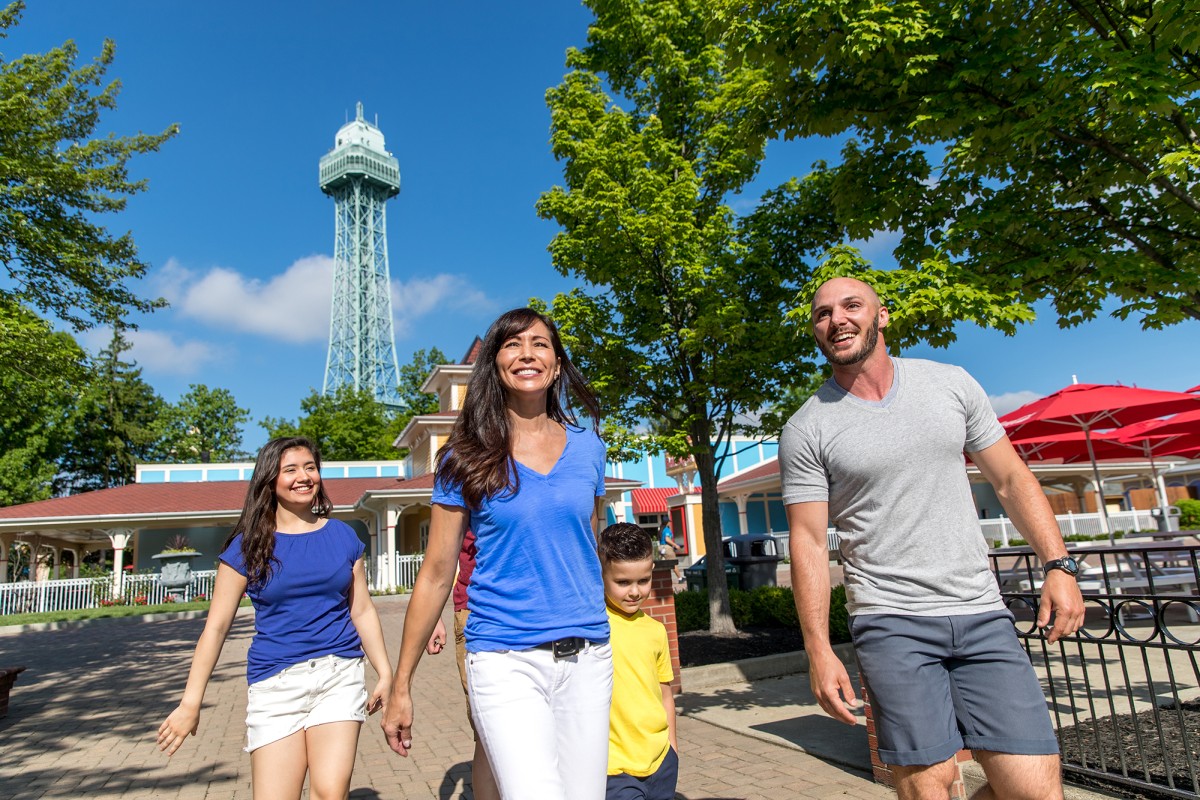 A family of four walking outdoors on a sunny day, with a tall lattice tower in the background and green trees around.