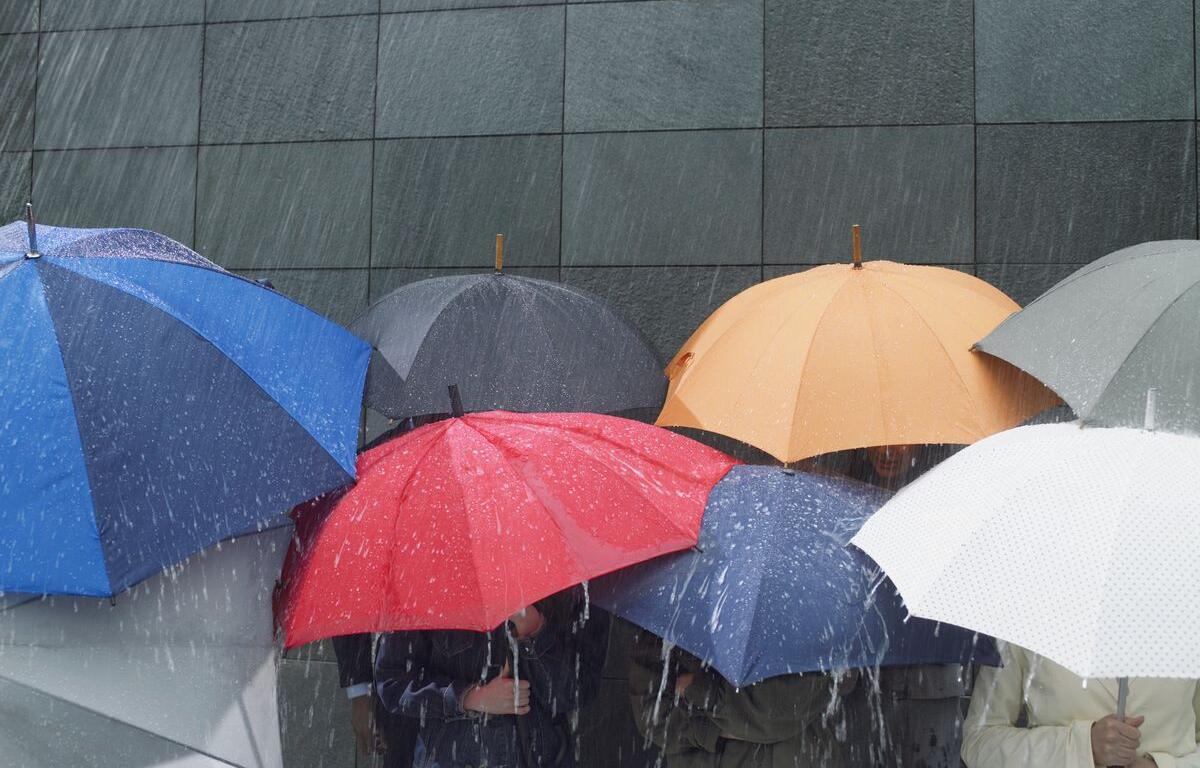 People holding colorful umbrellas in heavy rain against a dark tiled wall, partially obscuring figures beneath.