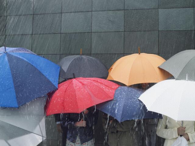 People holding colorful umbrellas in heavy rain against a dark tiled wall, partially obscuring figures beneath.