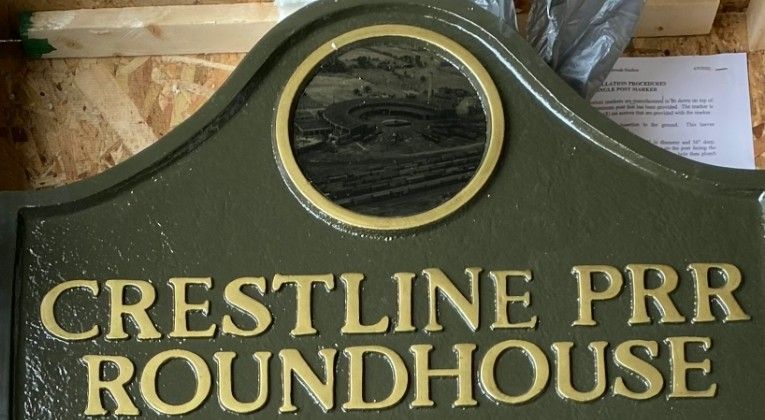 Green signboard with gold embossed lettering that reads 'CRESTLINE PRR ROUNDHOUSE' and a circular window at the top center, likely a railroad facility marker.
