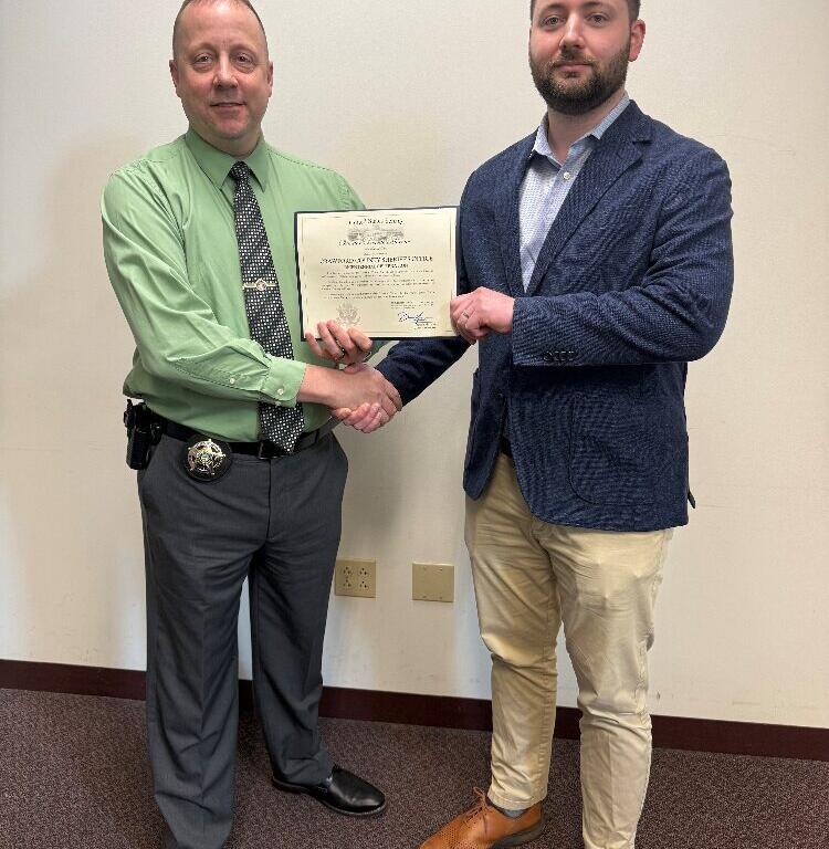 Two men shake hands while holding a certificate, one wearing a green shirt with a police-style badge, the other in a navy blazer and light shirt, standing in front of a plain wall.