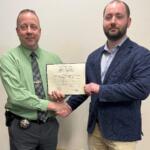 Two men shake hands while holding a certificate, one wearing a green shirt with a police-style badge, the other in a navy blazer and light shirt, standing in front of a plain wall.