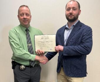 Two men shake hands while holding a certificate, one wearing a green shirt with a police-style badge, the other in a navy blazer and light shirt, standing in front of a plain wall.
