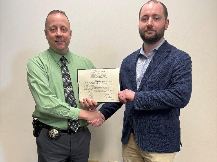 Two men shake hands while holding a certificate, one wearing a green shirt with a police-style badge, the other in a navy blazer and light shirt, standing in front of a plain wall.