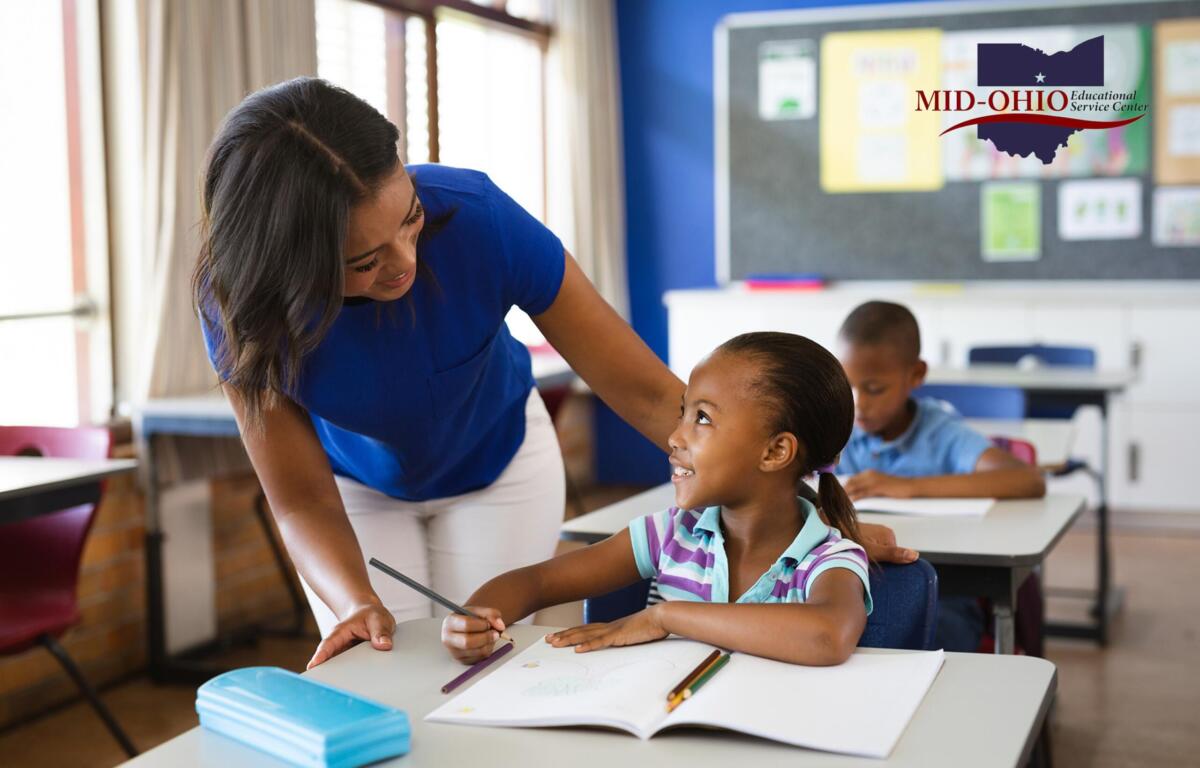 Teacher helps a smiling young girl with a pencil at a desk in a bright classroom; Mid-Ohio Educational Service Center logo visible on the wall.