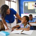 Teacher helps a smiling young girl with a pencil at a desk in a bright classroom; Mid-Ohio Educational Service Center logo visible on the wall.