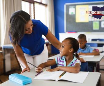 Teacher helps a smiling young girl with a pencil at a desk in a bright classroom; Mid-Ohio Educational Service Center logo visible on the wall.