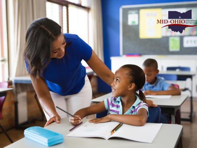 Teacher helps a smiling young girl with a pencil at a desk in a bright classroom; Mid-Ohio Educational Service Center logo visible on the wall.