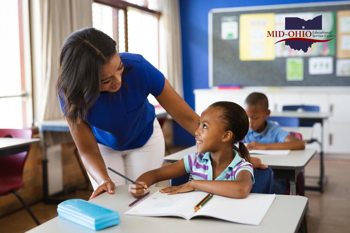 Teacher helps a smiling young girl with a pencil at a desk in a bright classroom; Mid-Ohio Educational Service Center logo visible on the wall.