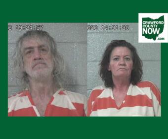 Two side-by-side mugshots: man with long gray hair and beard on the left, woman with dark hair on the right, both in orange-and-white striped jail uniforms against a gray cinder-block wall; timestamp overlays and Crawford County Now logo in corner.