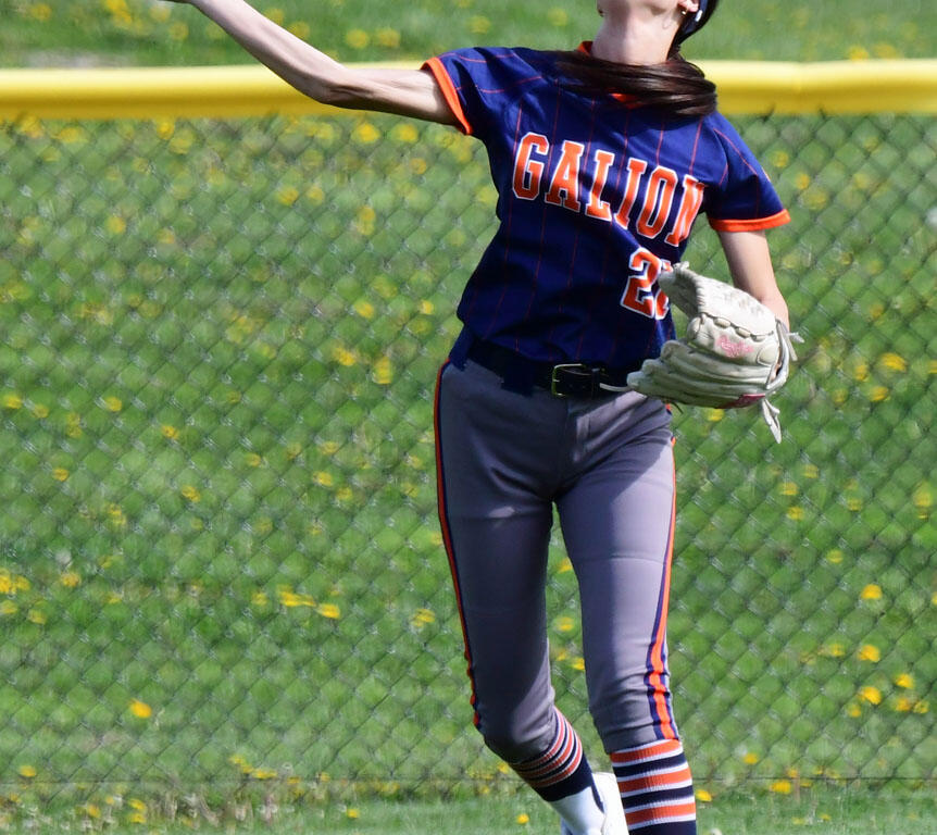 Softball pitcher in a blue and orange Galion jersey throws a bright neon-yellow ball on a grassy field behind a chain-link fence.