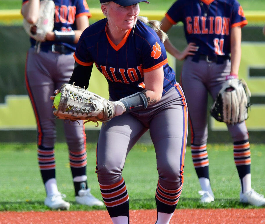 Softball player in a navy and orange Calion uniform crouches to field a ball with a glove, teammates in the background on a sunny field.