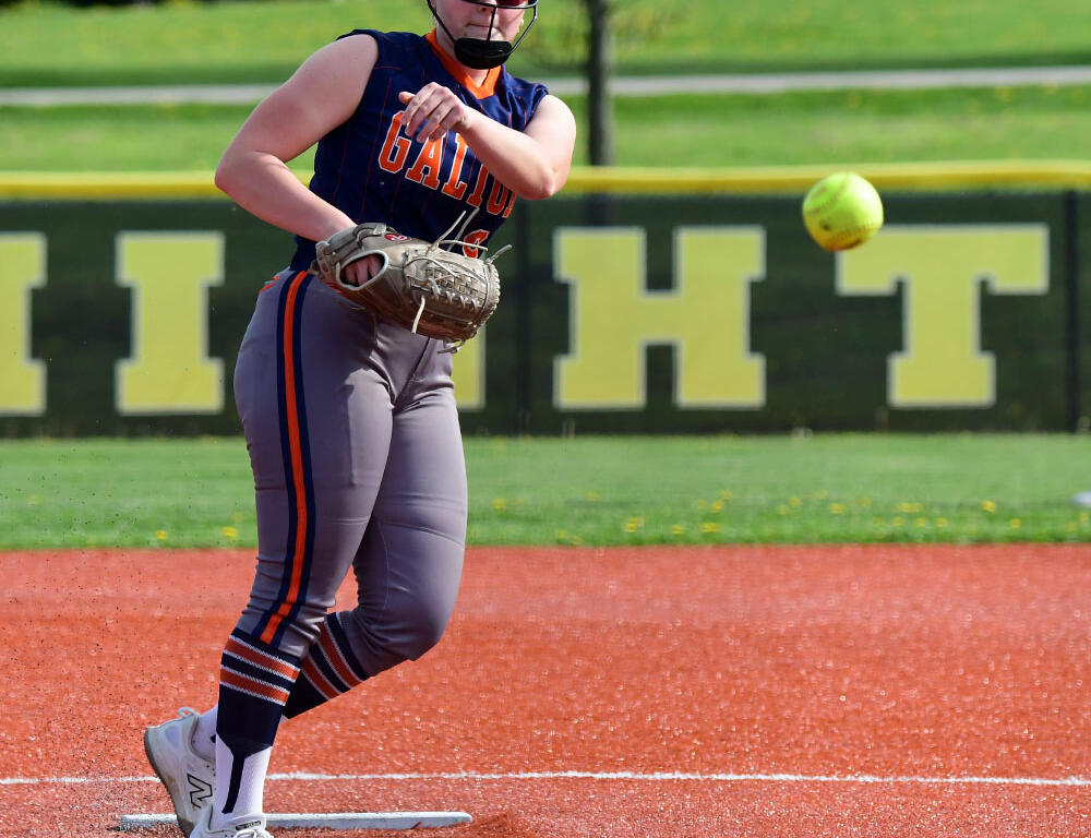 Female softball player on a base in a navy uniform with a glove on her left hand, ball approaching in the air near her