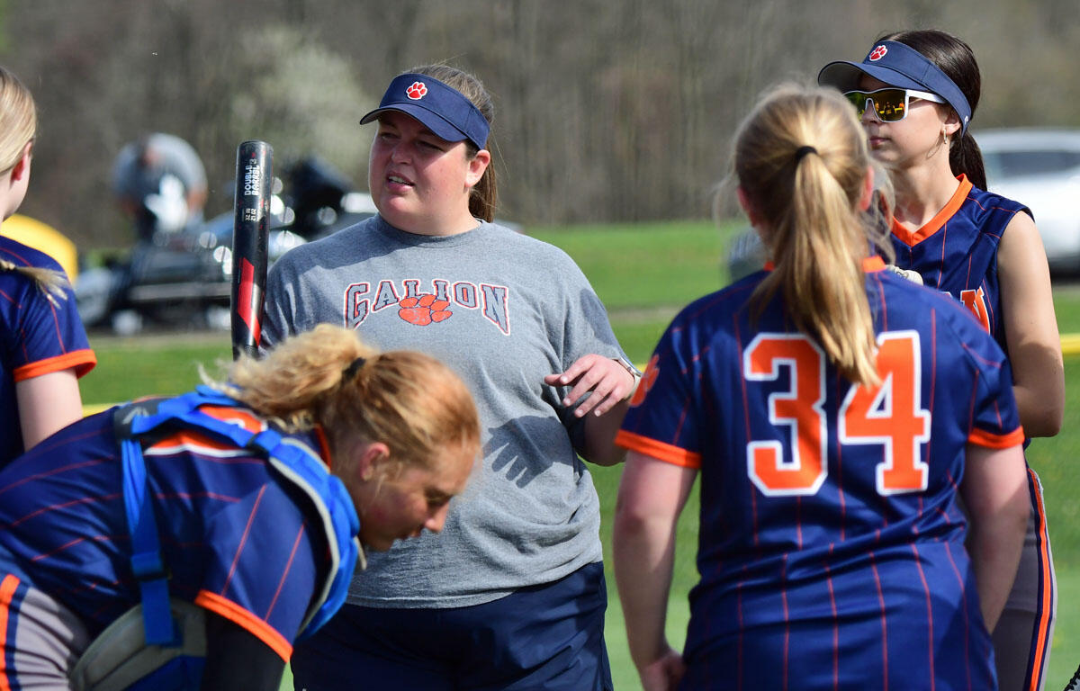 Softball coach in gray Gallion shirt speaks to players in blue-orange uniforms during a field practice on a sunny day, one holding a bat.