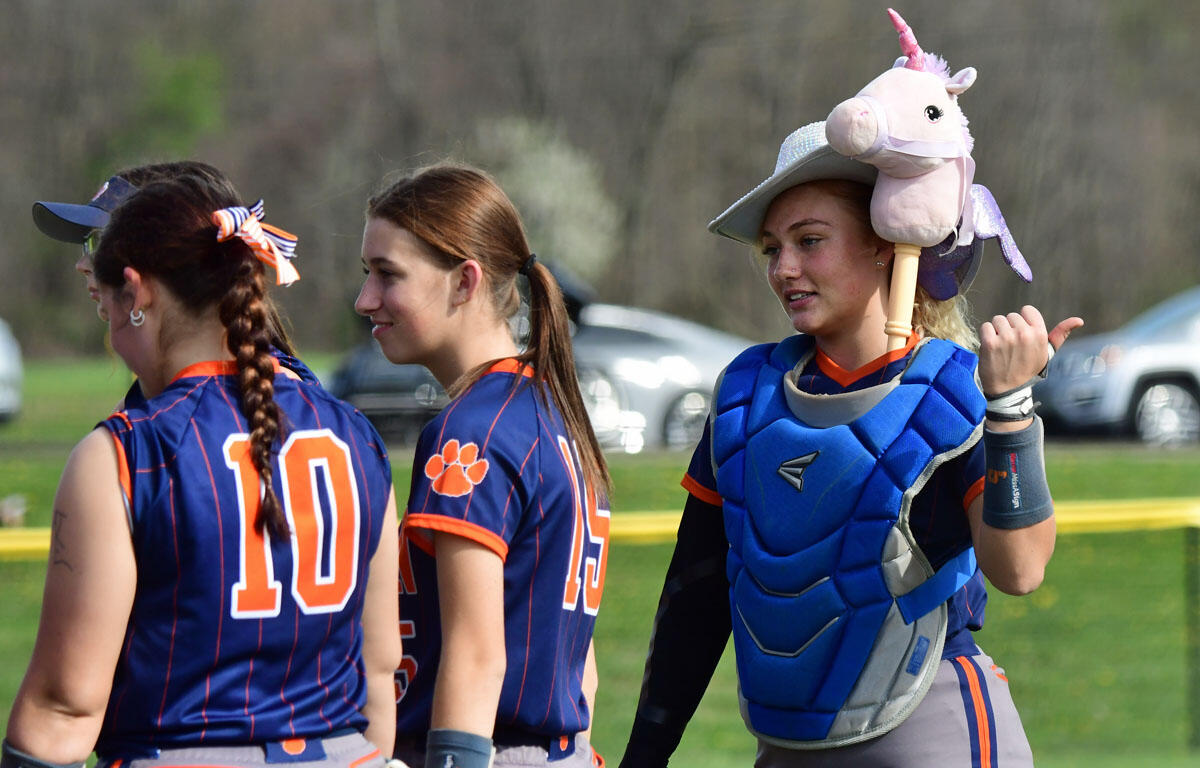 Three female softball players in navy uniforms with orange trim walk on a field; one wears a pink unicorn hat on a helmet and blue catcher's gear is visible on another player.