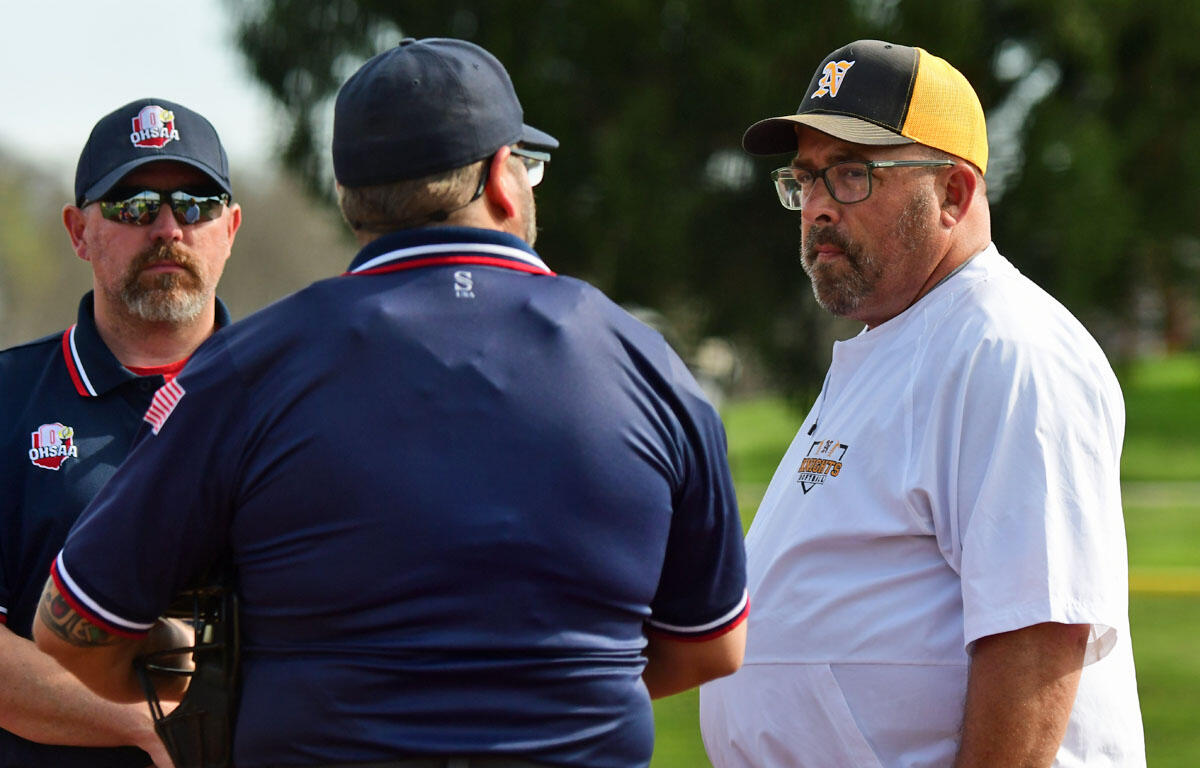 Three men wearing baseball caps and sports shirts stand in a sunny outdoor field, engaged in a serious discussion.