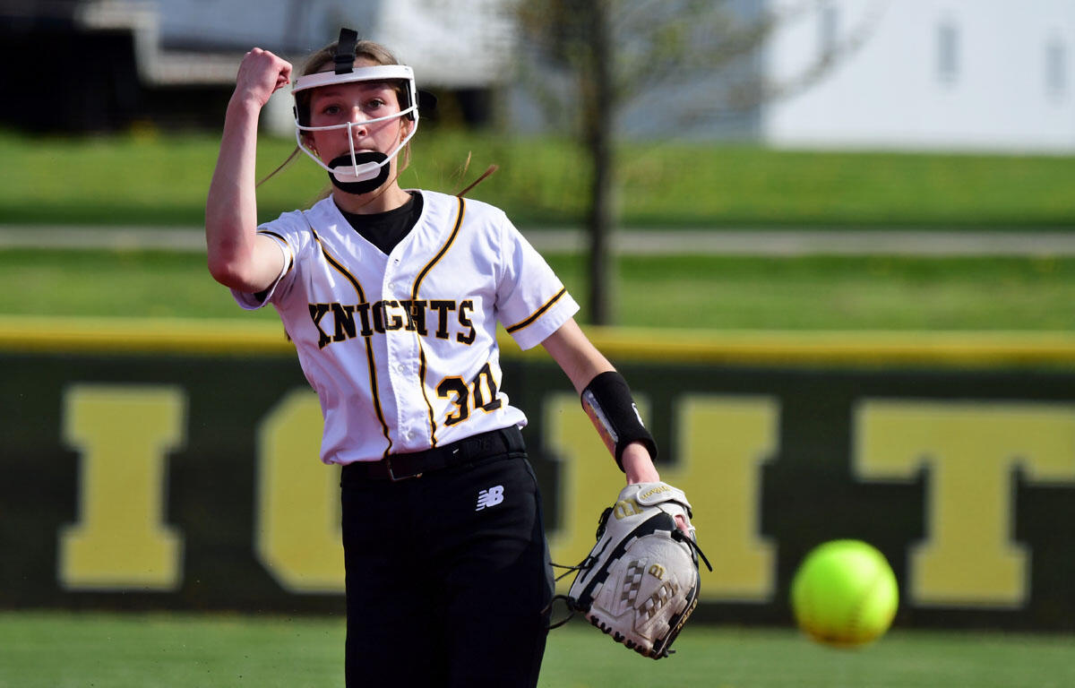 Female softball pitcher in a Knights white jersey prepares to throw a pitch on a sunny field, glove on her left hand and facing forward with a protective face mask helmet on.