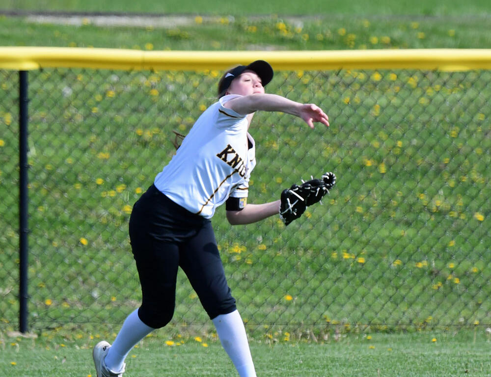 Softball pitcher in a white jersey and black cap throwing a ball during a game on a green field by a fence.