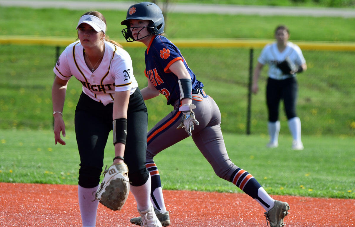 Two female softball players compete for a ball near a base: one in a white uniform with a glove crouches, the other in blue/orange with a helmet running after the ball.