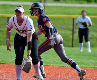 Two female softball players compete for a ball near a base: one in a white uniform with a glove crouches, the other in blue/orange with a helmet running after the ball.